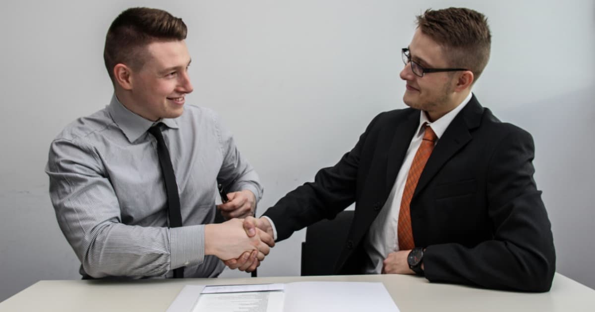 Two people shaking hands across a desk over a signed agreement after settling terms